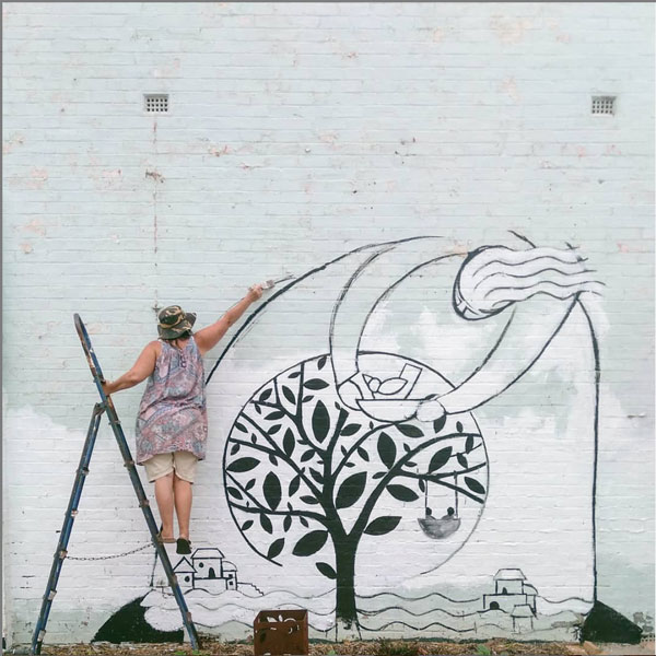 Mandy on a ladder painting a mural of the art garden logo 